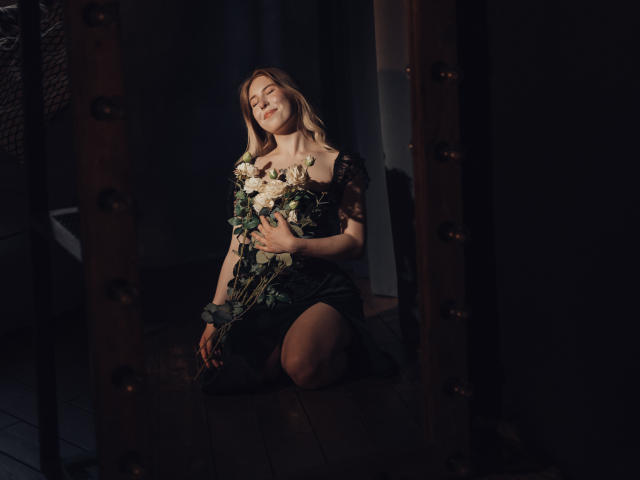 LanaRoselle kneeling in dim light, holding white and green flowers against her tits, wearing a black lace dress, with a soft, happy smile, and blonde hair.