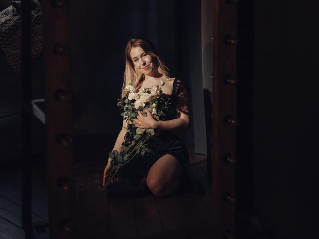 LanaRoselle kniet in einem dunklen Raum und hält einen Strauß aus weißen und grünen Blumen. Sie trägt ein schwarzes Spitzenkleid, wobei ihre Brüste leicht sichtbar sind, und lächelt sanft..