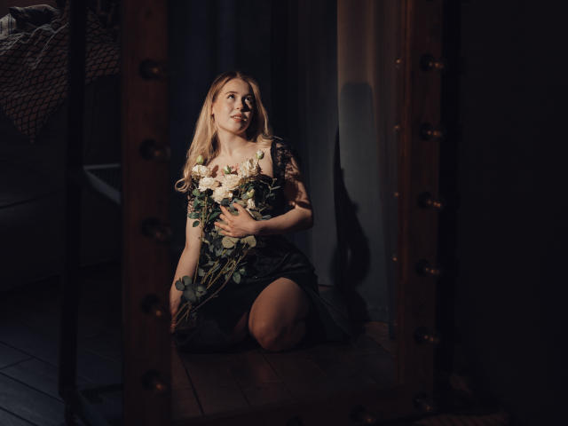LanaRoselle, rubia, arrodillada, sosteniendo flores blancas y verdes, vestido negro, mirando hacia arriba, con poca luz, marco de espejo, suelo de madera, sombras suaves, luz en su rostro, cabello ligeramente ondulado..