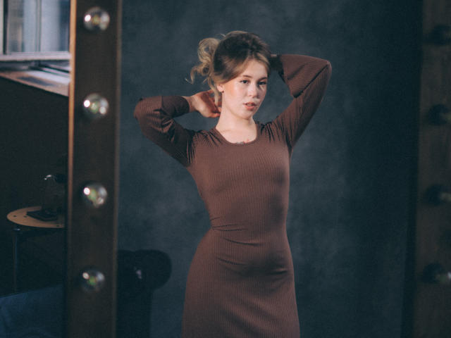 LanaRoselle, vêtue d'une robe marron côtelée, se tient debout, les mains derrière la tête, regardant sur le côté, dans une pièce faiblement éclairée, ses cheveux blonds en un chignon désordonné..