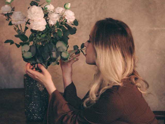 LanaRoselle mit blonden Haaren, in einem braunen Pullover, die weiße Rosen aus einer dunklen, glitzernden Vase riecht, eine Rose mit ihrer rechten Hand hält, Seitenansicht..