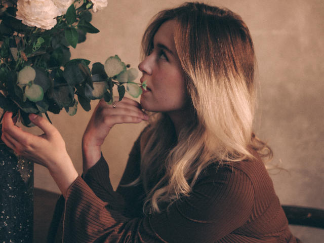 LanaRoselle with blonde, wavy hair, brown ribbed shirt, sniffing white roses and green eucalyptus leaves from a vase. She's looking to the side, holding her nose.