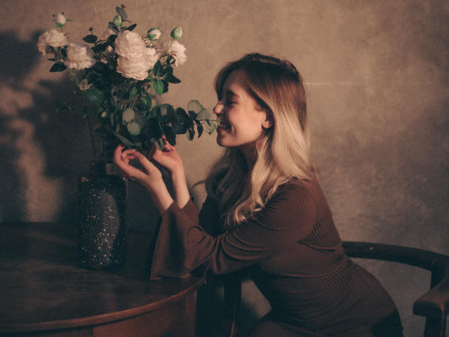 LanaRoselle met blond haar, lachend, gekleed in een bruin geribbeld topje, leunend op een tafel, met witte rozen in haar handen die uit een donkere vaas komen..