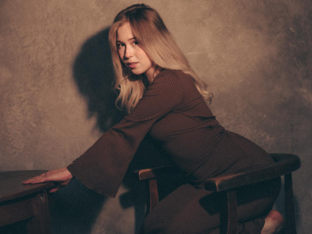 LanaRoselle, avec de longs cheveux blonds, une robe marron à rayures fines, appuyée sur une chaise en bois, regardant par-dessus son épaule, lumière douce, ton chaud, ombre subtile, expression douce, petite fesse, silhouette mince..