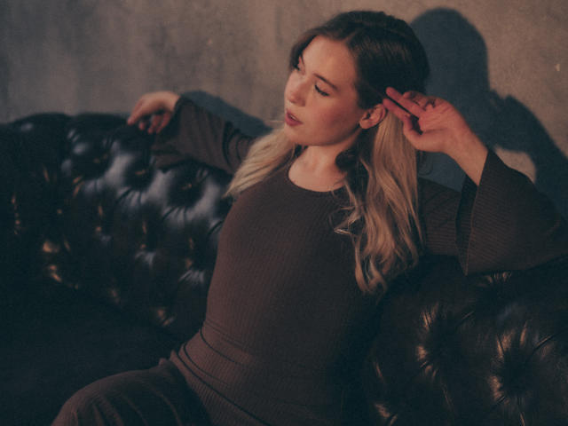 LanaRoselle met blond haar, gekleed in een bruine gebreide trui, leunt tegen een donkere leren bank met knopen, haar hand in haar haar, zacht licht, ontspannen, licht gedraaid, melancholische uitdrukking..