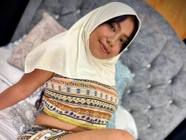 AliKayleigh with dark hair, wearing a white hijab and a patterned top, sitting on a bed, smiling, showing a small ass, and wearing colorful, patterned shorts.