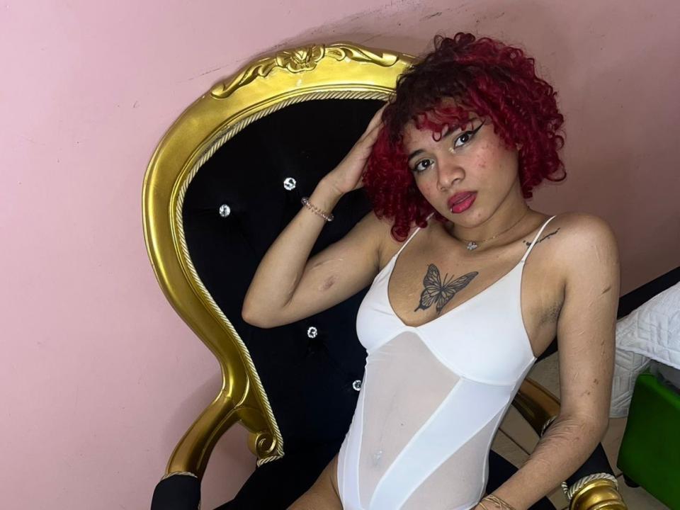 CaroliineFoxy mit roten, lockigen Haaren, weißem, durchsichtigen Body, Schmetterlingstattoo auf der Brust, Hand im Haar, sitzend auf einem schwarzen und goldenen Stuhl, rosa Wand, leicht verschmutzte Haut..