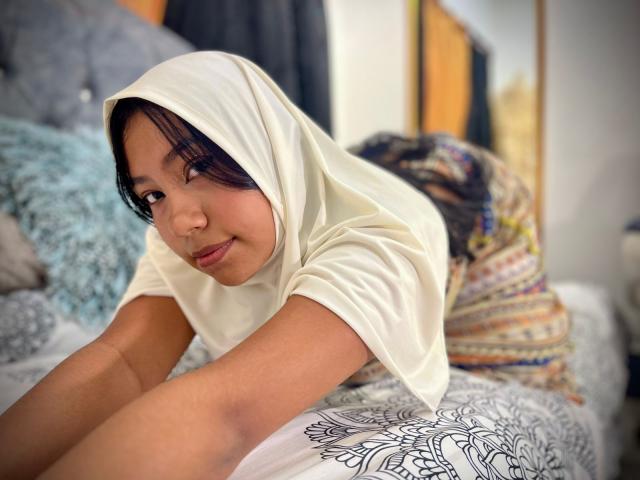 AliKayleigh is on a bed, wearing a white headscarf and white shirt, leaning forward with a cute, slightly smug expression, showing her ass and dark, messy hair.