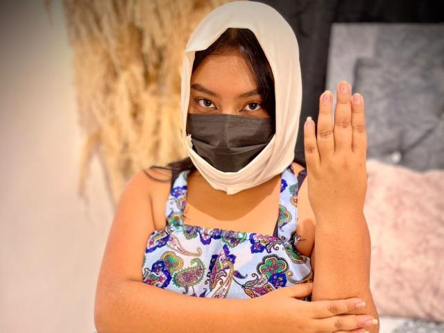AliKayleigh with a white hijab, black mask, and colorful dress, holding her foot up. She's got brown eyes and tan skin, looking at the camera. Her foot's up in the air.