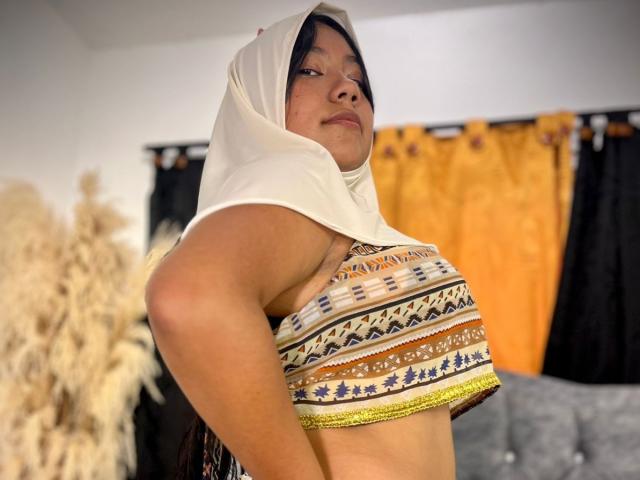 AliKayleigh with a white headscarf, wearing a patterned, sleeveless top that exposes her small, firm tits, looking slightly to the side with a confident expression.