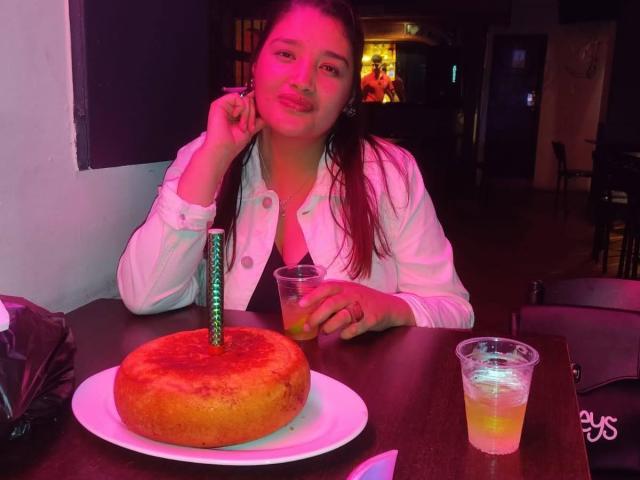 ZafiRouge with pink glow, wearing white shirt, smiling, holding drink, cake with green candle on plate, glass of yellow drink, in dimly lit room.