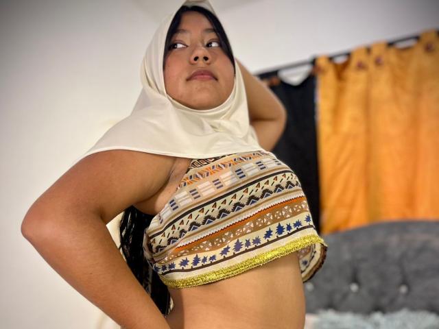 AliKayleigh has a chubby face, dark long hair, and wears a white headscarf with a patterned tube top showing her small boobs. She's looking up with a hand on her head.