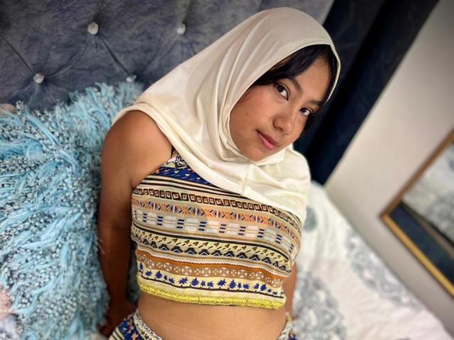 AliKayleigh has tan skin, dark hair, and wears a white headscarf. She's in a patterned crop top, sitting on a bed with blue and white pillows. Boobs are visible.