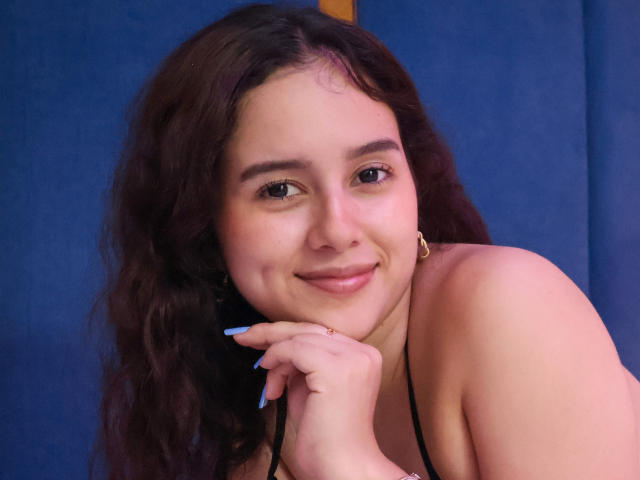 EmmieBellefort with wavy brown hair, blue nails, smiling softly, black spaghetti strap top, resting chin on hand, blue background, cute, medium breasts, subtle makeup, gold earrings.