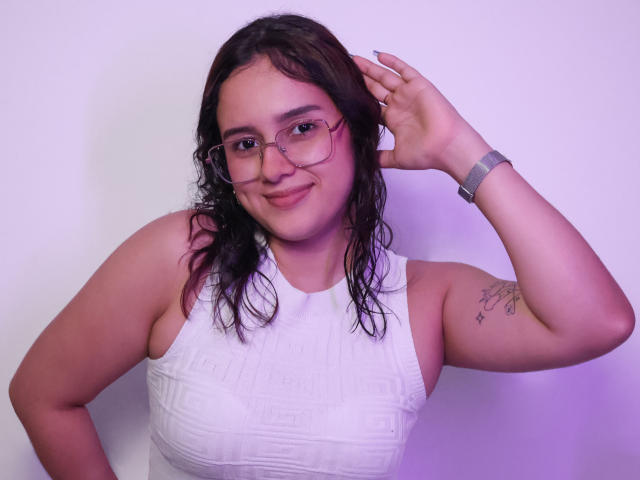 EmmieBellefort with wavy brown hair, glasses, white textured dress, tattoo on right arm, smiling, hand in hair, wearing a grey watch, light skin, slight purple lighting.