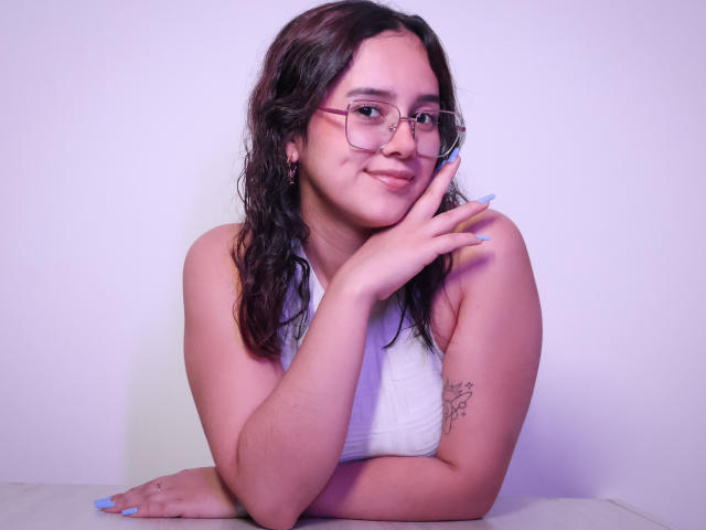EmmieBellefort with wavy brown hair, glasses, white sleeveless top, smiling, finger on cheek, tattoo on right arm, blue nails, leaning on white surface, looking cute and sexy.
