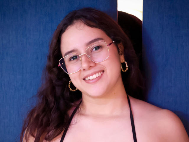 Emmie Bellefort, met krullend bruin haar, een bril en gouden oorringen, lacht met witte tanden, draagt een zwarte haltertop, en de achtergrond is blauw met natuurlijk licht. Haar borsten zijn licht zichtbaar..
