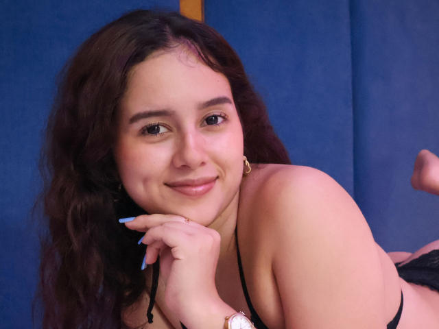 EmmieBellefort has wavy brown hair, light skin, and a cute smile, lying down with black lingerie, showing her small tits, and blue nails, against a blue background.