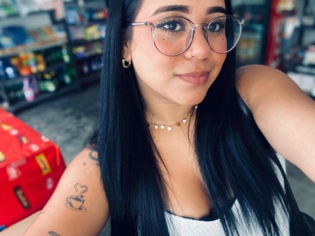 AuroraDiMarco with long black hair, glasses, and a small nose piercing, wearing a white top, showing a tattoo of a cupcake on her arm, and a necklace.