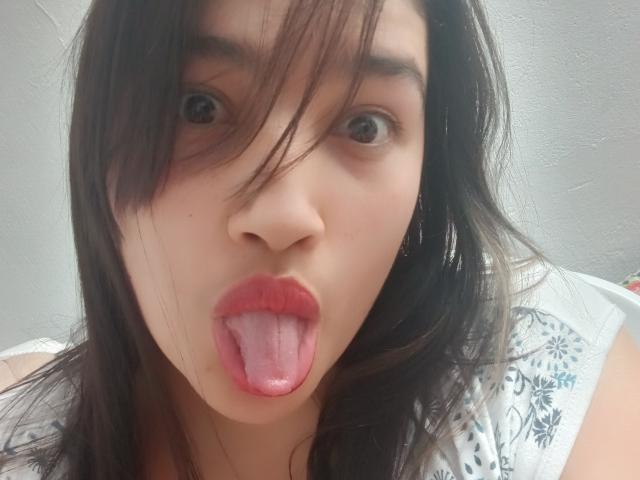 ZafiRouge has dark brown hair, wide brown eyes, and is sticking out her pink tongue with red lipstick, wearing a white shirt with blue floral patterns.