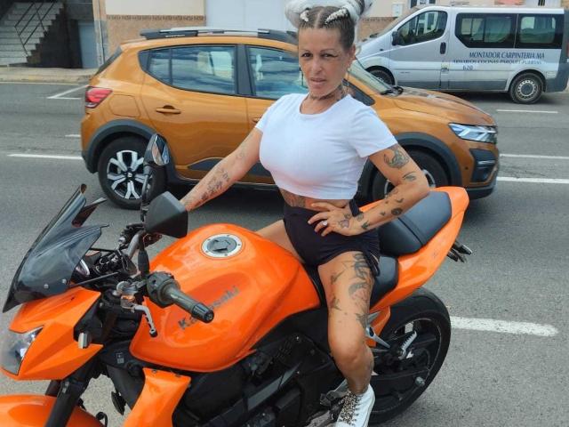 CanariaHot with tattoos, white crop top, black shorts, and grey sneakers, sitting on an orange motorcycle, hands on hips, in an urban street, with a yellow car behind.