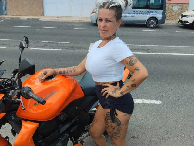 CanariaHot with tattoos, white crop top, black shorts, standing next to an orange motorcycle, hands on hip, street in background, white van with text in background.