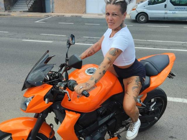 CanariaHot with tattoos, white crop top, black shorts, and white sneakers, riding an orange motorcycle, holding the handlebars, smiling.