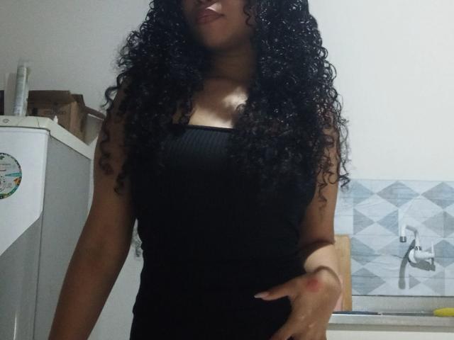 Lumminah with long curly black hair, wearing a black ribbed tank top, standing in a kitchen, hand on her hip, showing her tits, medium brown skin, subtle smile.