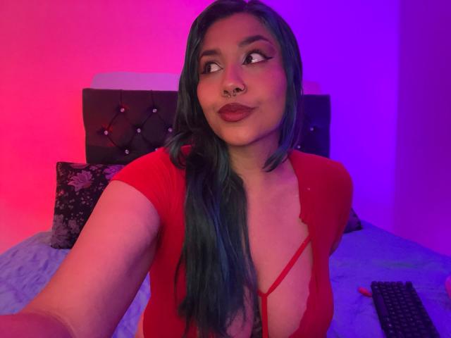 LilithHela with green hair, red lipstick, and nose ring, wearing a red, low-cut top, showing her tits, in a purple and pink-lit bedroom, taking a selfie.