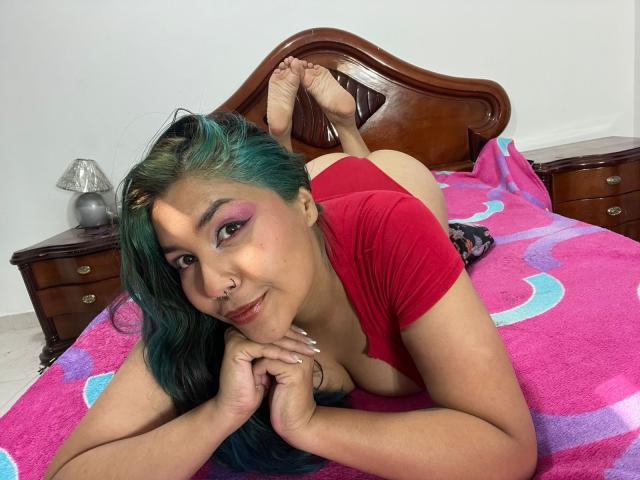 LilithHela with teal-green hair, red top, and pink eyeshadow, lying on a pink bed, butt up, legs crossed, smiling, hands under chin, showing tits and ass.