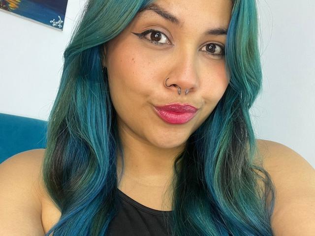 LilithHela with teal and blue hair, nose ring, and pink lipstick, pouting with makeup accentuating her eyes, wearing a black top, and looking into the camera.