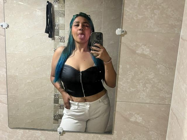 LilithHela with blue hair, tongue out, wearing a black leather strap top and white pants, taking a mirror selfie with her right hand in a tiled bathroom.