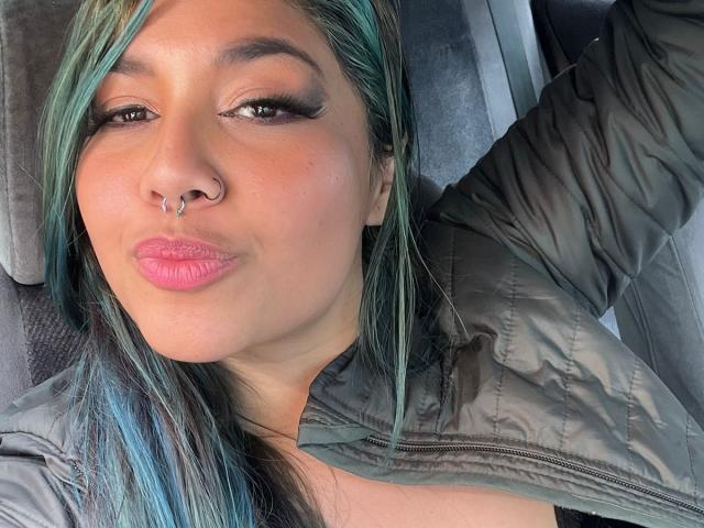 LilithHela with teal and blue hair, pouty pink lips, nose ring, and green jacket, lying down, looking at camera. Her arm is raised. Her tits are slightly visible.