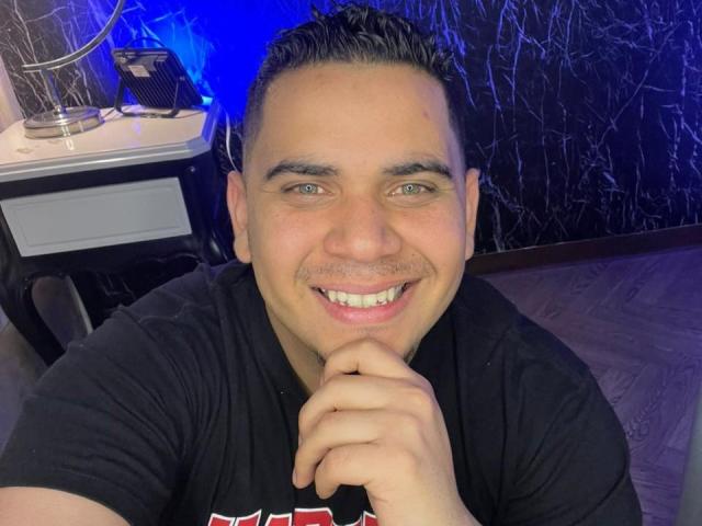 DmitriIvanov with short, dark, slicked-back hair, smiling, green eyes, black shirt, hand under chin, blue and black background, and a small white desk with a lamp.