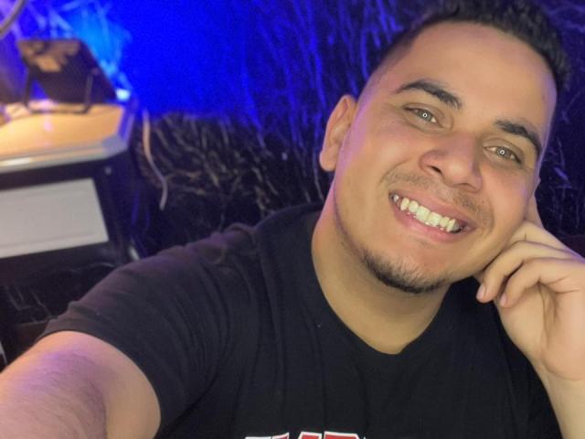DmitriIvanov with a smiling face, short dark hair, light skin, black shirt, and a cute goatee, resting his cheek on his hand in a selfie. Blue light in background.