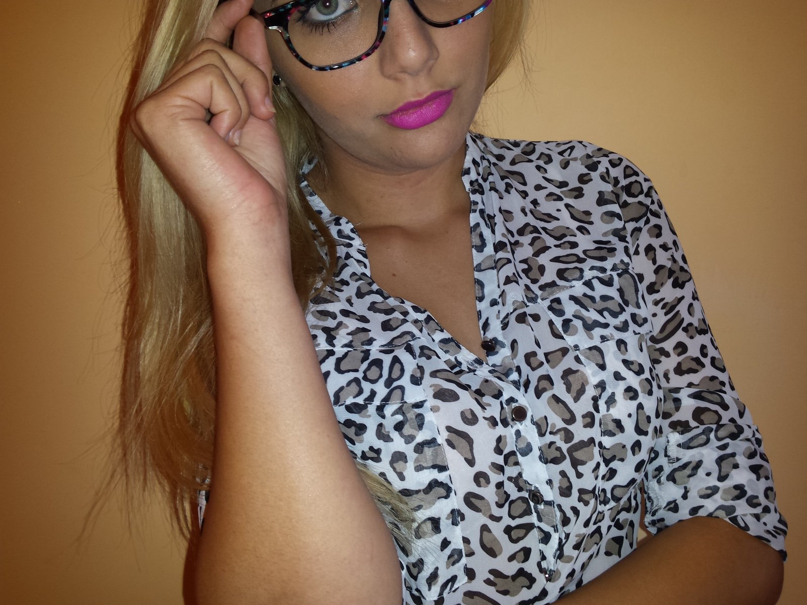 NastySabrynna, com cabelo loiro, usando óculos e uma camisa de estampa de leopardo, tem batom rosa e está tocando sua orelha direita com a mão. Os seios estão parcialmente visíveis..