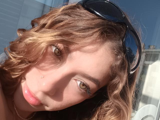 El rostro de TatianaVida, con cabello castaño ondulado, ojos verdes, labios rosados, un pequeño piercing en la nariz y gafas de sol negras en la cabeza. La luz del sol resalta su piel..