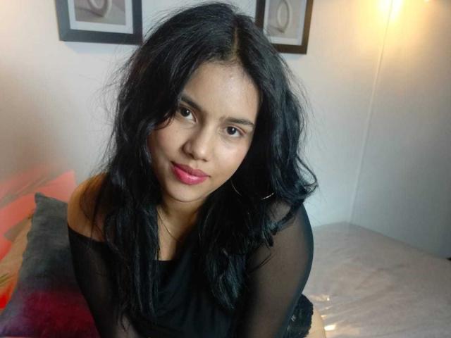 Allise has dark wavy hair, brown eyes, pink lipstick, and is wearing a black sheer top, sitting on a bed with red and gray pillows.