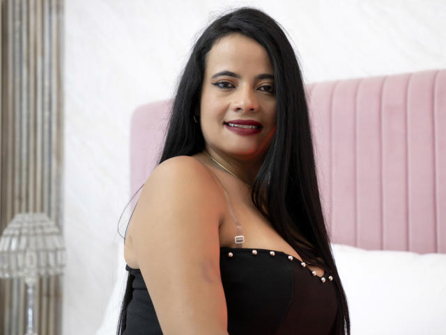 Paola Valencia ha i capelli neri, un seno di media grandezza, e sta sorridendo con il rossetto rosso, indossando una maglietta nera con piccoli bottoni dorati, in piedi di fronte a una testiera rosa..