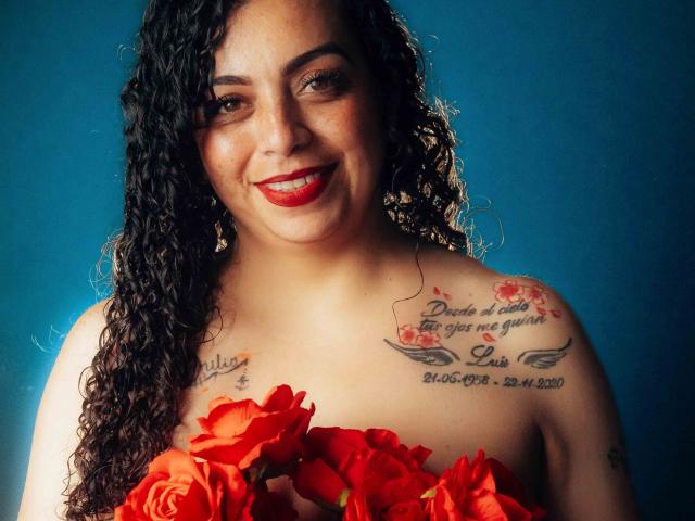 AlissaaMilf with curly black hair, red lipstick, holding red roses, smiling at camera, has tattoos on both shoulders, blue background.