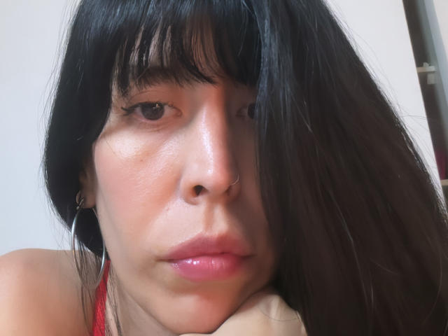 SeleneBrigth has shiny black hair, bangs, hoop earrings, nose ring, glossy pink lips, and a slight pout, resting her chin on her hand. Her skin is smooth and tan.