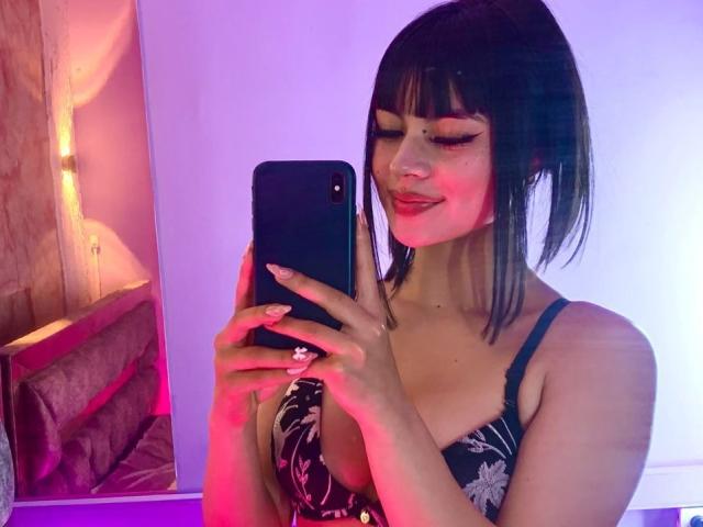 PinkSuni with black hair in a ponytail, wearing a floral black dress, holding a phone, smiling, showing off her tits, pink and purple lighting, pink nail polish.