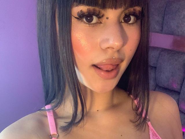 PinkSuni's face, brown straight hair, pink straps, tongue out, small nose piercing, freckles, shiny pink cheeks, big brown eyes with long lashes, slightly open mouth.