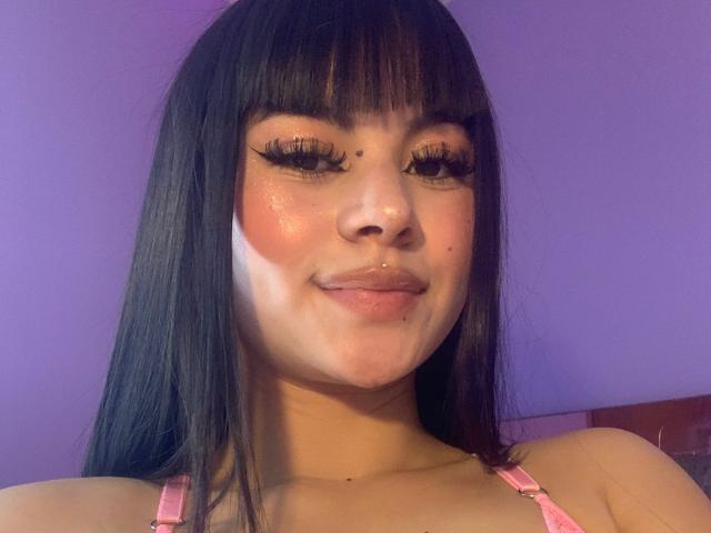 PinkSuni with long black hair, wearing a pink bra, has shiny skin, dark eyeliner, and a small black mole above her right eyebrow. Her face is slightly messy.