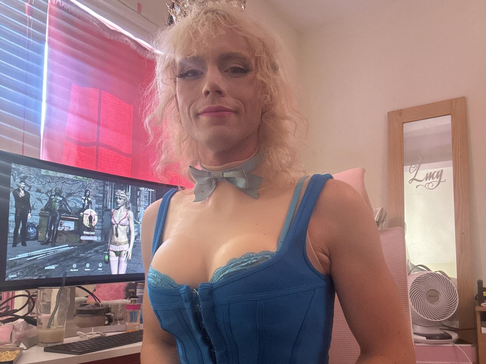 KaliTGirl con i capelli biondi ricci, un corpetto blu con lacci, che mostra il seno, indossa una collana blu con un fiocco, in piedi di fronte a un computer con un programma televisivo sullo schermo..