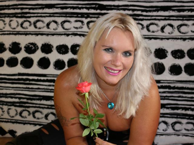 LadyMargaret-hot with blonde hair, blue eyes, pink lipstick, holding a red rose, wearing a blue gem necklace, showing her tits, smiling, in a black and white patterned backdrop.