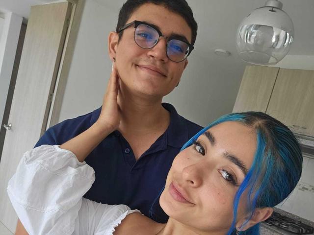 ConnieXMarc with blue hair and white top, smiling, stands next to ConnieXMarc with glasses and dark shirt, hand on neck, in a room with a hanging light.