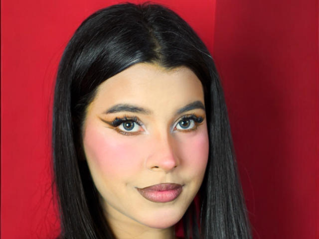 ArtemisaStone has black hair, green eyes, pink blush, winged eyeliner, and pink lipstick, with a subtle smile, against a red background. Her tits are not visible.