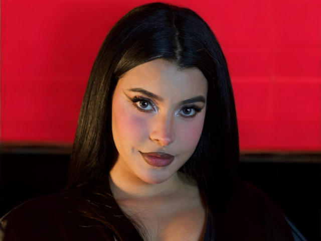 ArtemisaStone with long black hair, brown eyes, and pink cheeks, wearing dark lipstick, looking at the camera with a subtle smile, against a red background. Boobs visible, cleavage.