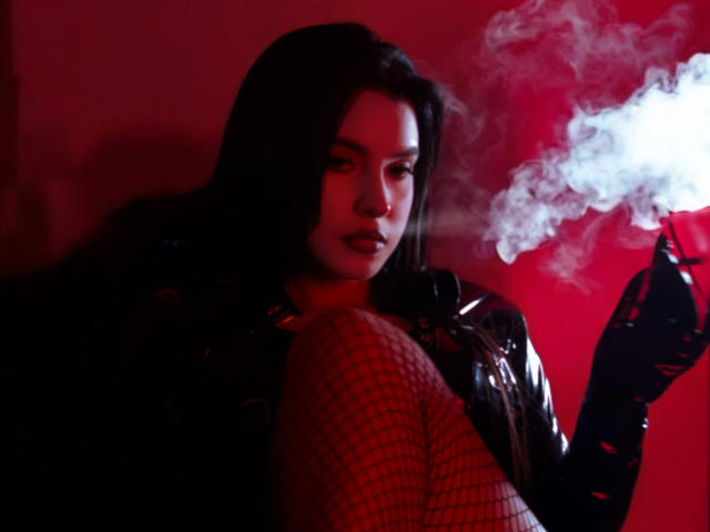 ArtemisaStone with dark hair, red-tinted face, holding a smoking cigarette, wearing a shiny black jacket and red fishnet leggings, looking sexy and sultry.